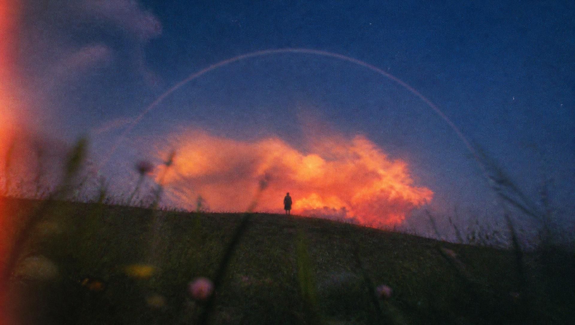 A lone figure beneath a glowing cloud and faint arc at twilight